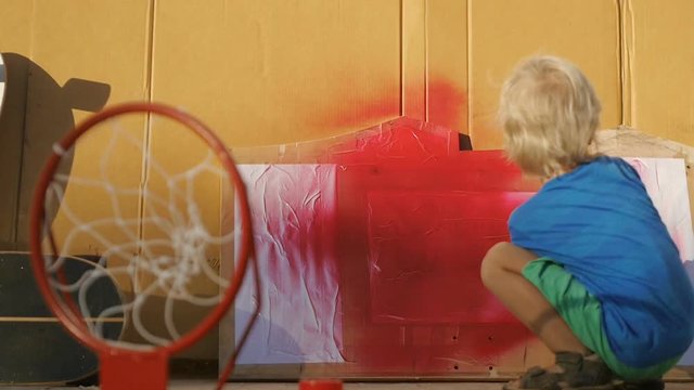 Little adorable child painting by spray a wooden board for basketball hoop by himself. Family working together in a workshop enjoying summer holidays.