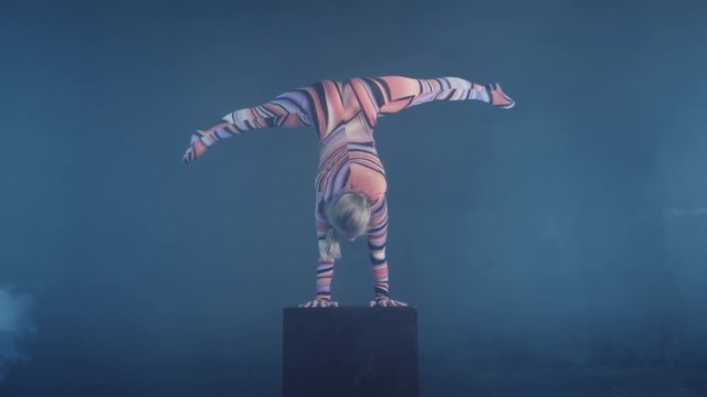 Young flexible blonde circus acrobat posing in studio in costume. Doing equilibre balance handstand on a cube. Copy space text.