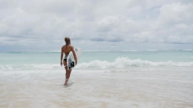 Slowmotion footage of a girl walking at the beach with her surfboard.
