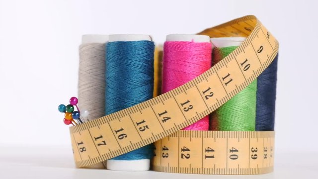 Set of colorful spools coiled by yellow measuring tape, pins with multicolor heads and needle with thimble on white background, sewing, equipment for tailors. Close up, seamless loop 4K Ultra HD.