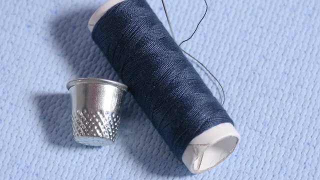 Dark blue bobbin, silver thimble and needle sticking out from piece of blue cloth, isolated tools for sewing, haberdashery. Close up, top view, seamless loop rotation, 4K Ultra HD.