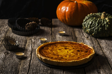 American pumpkin pie on a dark shabby wooden background with candles, baking dishes and fir cones. Dark photo. Selective focus 