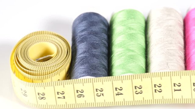 Bobbins with threads in row, measuring tape, pins and needles, silver thimble and small buttons lay on white surface, equipment for tailoring, haberdashery. Close up, 4K Ultra HD.