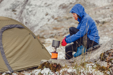 Woman boiling water on portable camping gas-jet near the hiking tent in snowy late autumn mountains