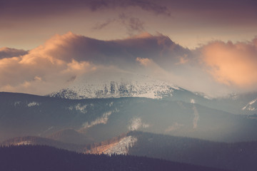 Landscape of winter mountains at sunset: snow covers  hills and  wooded peaks. Dramatic evening sky. 