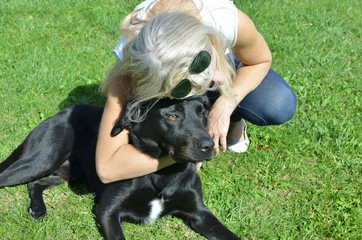 Woman hugging her adorable black dog who is lying on a green lawn