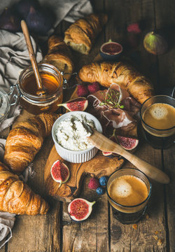 Breakfast with croissants, ricotta cheese, figs, fresh berries, prosciutto meat, honey in glass jar and espresso coffee over rustic wooden background, selective focus, vertical composition