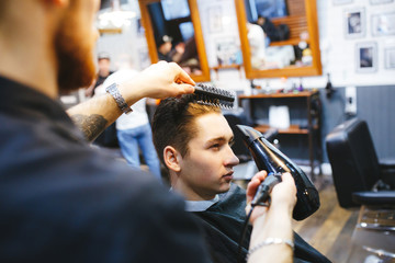 Male barber makes hair styling using a dryer.