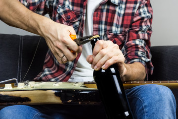 Opening beer with pliers. Male person in casual wear changing strings on electric guitar opens a bottle of drink with tool to have a break