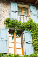 Fototapeta na wymiar Window with shutters in Mediterranean ambience. Cheerful summer colors. Green plants climbing the wall.