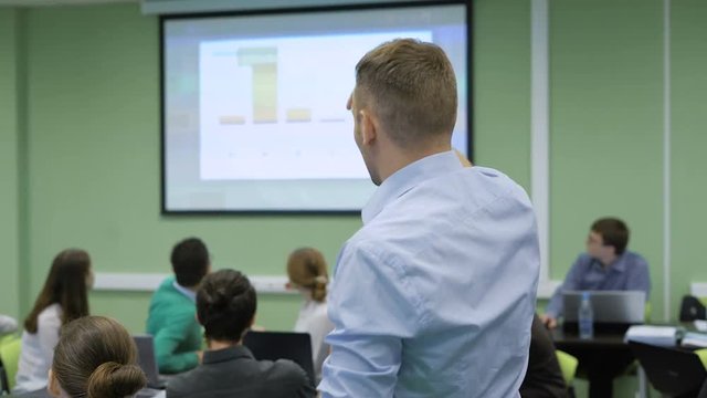 Professional teacher in class room at the university is delivering lecture on Economics pointing highlights and schemes on the screen on the wall for students on Finance Faculty.