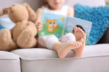 Funny little girl reading book on sofa at home
