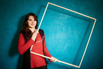brunette woman in a red blouse standing against the backdrop of a blue wall and holding a large wooden frame