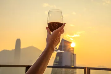 Foto op Plexiglas Wijn Close up of glass of red wine raised with the background the spectacular Hong Kong skyline at sunset. Rooftop drinks overlooking the city skyline.