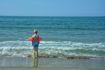 Kleiner blonder Junge mit roten Schwimmfl&uuml;gel steht iam Strand  vor einer Welle und schaut zum  Meer
