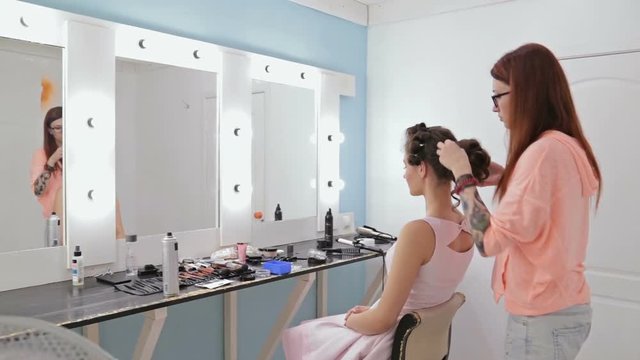 Top knot hair design - hair stylist, hairdresser in glasses finishing hairstyle for young pretty woman and using bobby pins for fixing hair in white make up room. Beauty and haircare concept
