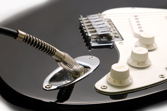 Part of modern electric six string guitar black color with glossy finish with cord in socket, pickups and control knobs isolated on white background horizontal view close up