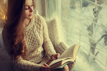 Young beautiful girl sitting on windowsill, holding and reading book. Model wearing classic stylish knitted dress. Day light. Christmas, New year, winter holidays concept. Copy space for text. Toned