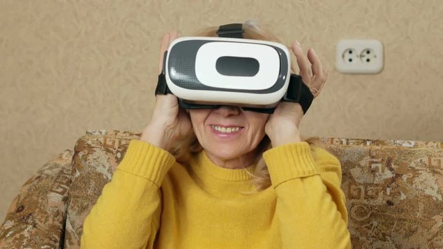 Senior woman wears glasses of virtual reality and watching a movie. She looks around and wants to touch the virtual walls. holiday home concept