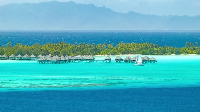 Bora Bora Bungalows Over The Tropical Shades of Blue Water and a Passing Sailboat in the Exotic French Polynesia Island of the South Pacific