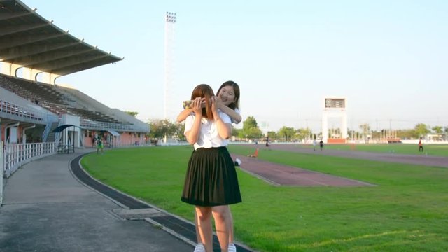 Cute Asian Thai college student girl in university uniform playing with her mobile and another girl come and close her eyes with surprise and hugging each other in loving friendship leisure concept.