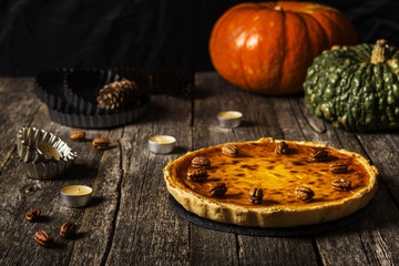 American pumpkin pie on a dark shabby wooden background with pecans, candles, baking dishes and fir cones. Dark photo. Selective focus 
