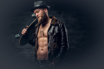 Bearded male dressed in leather jackett holds skateboard.