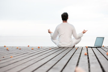 Man in white clothes meditating yoga with laptop on wooden pier
