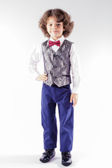Cute curly boy in a silver waistcoat is facing the camera and looking into the camera. Full length. Gray background.