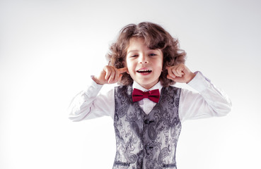 Curly cute boy in waistcoat and bow tie laughing closing ears with your fingers. Close-up. Gray...