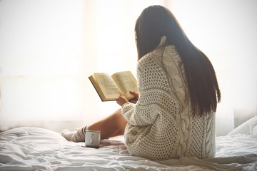 Young woman sitting in bed while reading a book - obrazy, fototapety, plakaty