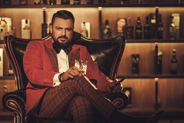 Extravagant stylish man with whisky glass sitting on armchair in gentleman club