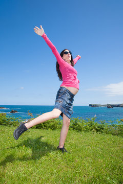 brunette brown pregnant woman with pink jersey blue jeans skirt and black sunglasses happy dancing on green grass meadow next to ocean
