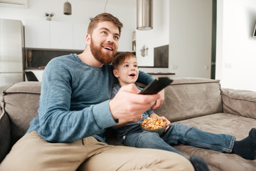 Happy father watching TV with his little son holding popcorn.