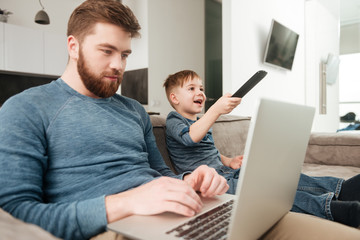 Bearded father using laptop computer while his son watching TV.