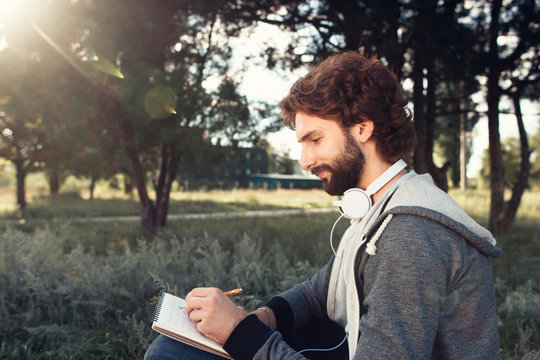 Young man writing in notebook at nature, side view. Profile of bearded man creating composition, free space for text. Art, idea, inspiration concept