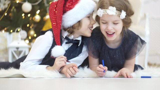 Children write letters to Santa Claus. To children it is cheerful. Little boy and girl lie on a floor near Christmas tree. House is festively decorated. Under Christmas tree there are a lot of gifts.