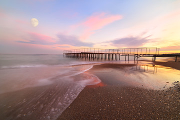Evening on the sea, a gentle sunset sky, waves run ashore, old pier. Delicate shades of evening. Large moon in the sky. Soft focus. Long exposure. The water motion blur.
