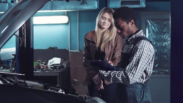 Mechanic discussing a car service with the attractive blond female owner standing writing on a job card viewed across the open bonnet and engine