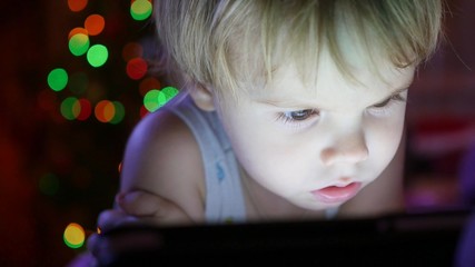 Fototapeta na wymiar The child looks to the tablet lying on bed. In the background, lights and garlands of Christmas fir