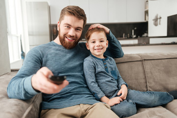 Father holding remote control while watching TV with little son.