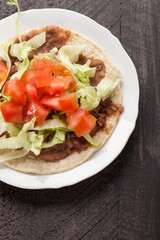 Corn tortilla tostada with refried beans, lettuce, salsa, and tomatoes top view on a dark wooden background