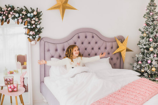 Portrait of cute funny little girl on Christmas morning in white home interior. Child cheerfully sitting in bed. Horizontal color image