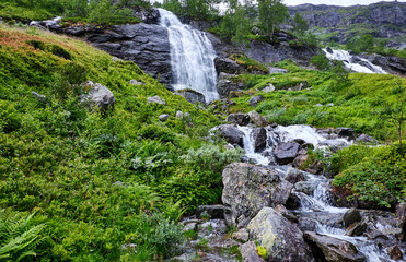 Fototapeta na wymiar Green and luxuriant nature in Norway, with gushing water in waterfalls and calmly running water on terraced boulders, beneath the high mountain ridge