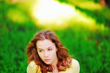 Fototapeta premium Portrait of young beautiful pensive girl brown-haired women with stunning eyes posing in the Park a summer day at blurry background of bright green lush grass with a beam of sunlight, closeup.