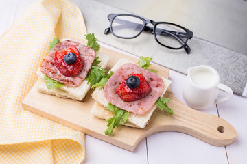 Healthy breakfast with toasted bread, milk, ham, vegetables, strawberry, jam and blueberry topping on wooden plate. Top view with white wooden table background.