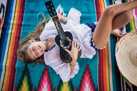 young girl plays ukulele while laying on a colorful Mexican blanket
