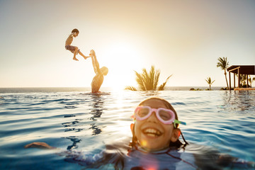Father throws son in the pool with daughter in forground wearing goggles and laughing