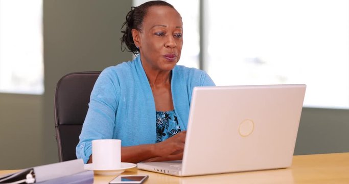 A black businesswoman happily uses her laptop at her desk. An African American woman works on her computer