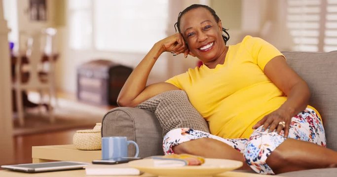 An older black woman is very happy as she poses for a  portrait while sitting on her couch. An elderly African American woman looks at the camera cheerfully